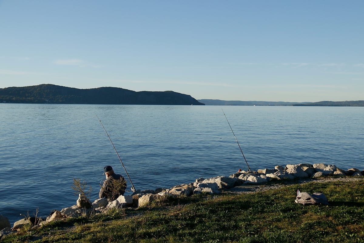 A man fishing from the grassy shore, two rods against the rocks, the river and hills beyond.