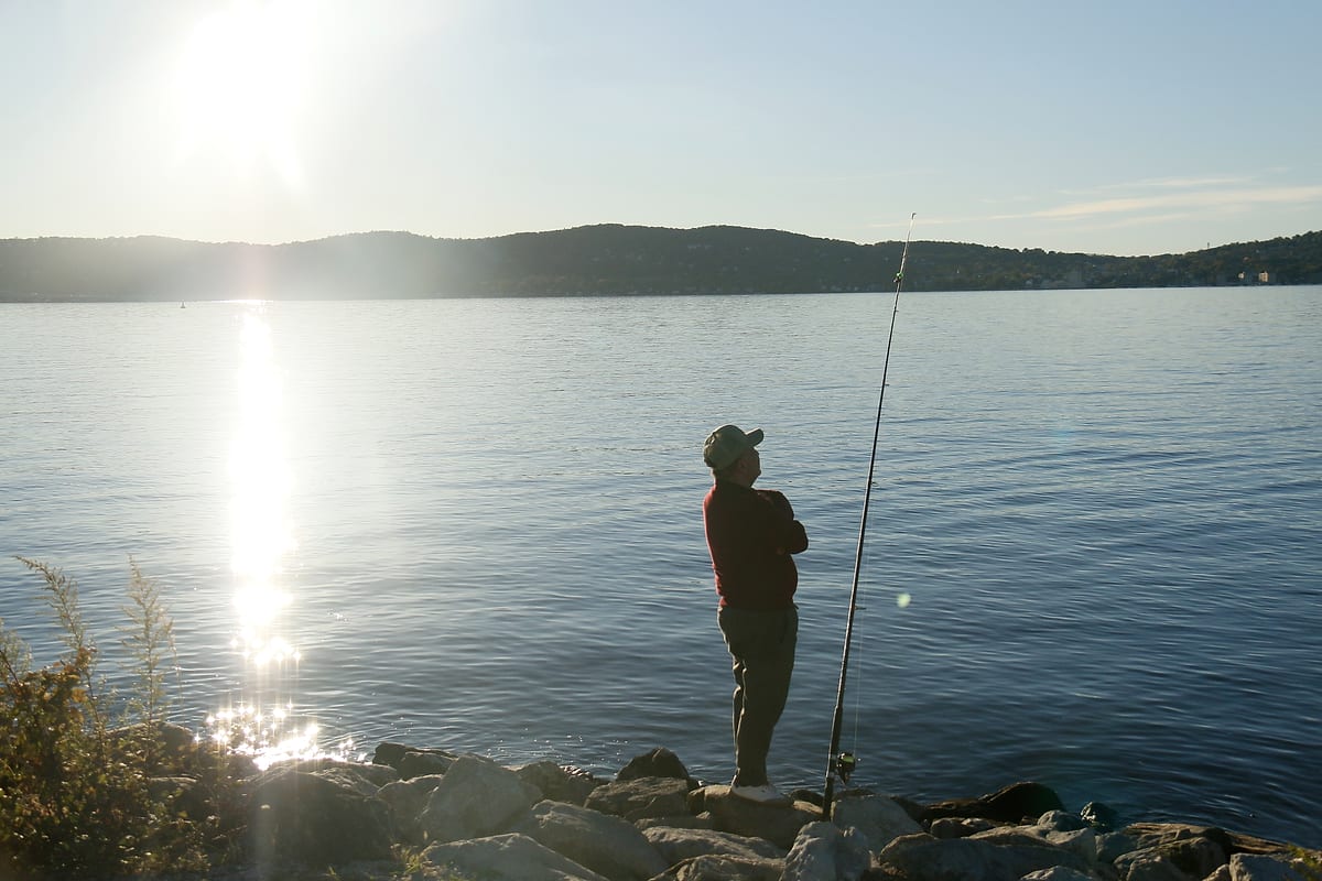 A man fishing from the rocks looks up, the sun's reflection beside him.