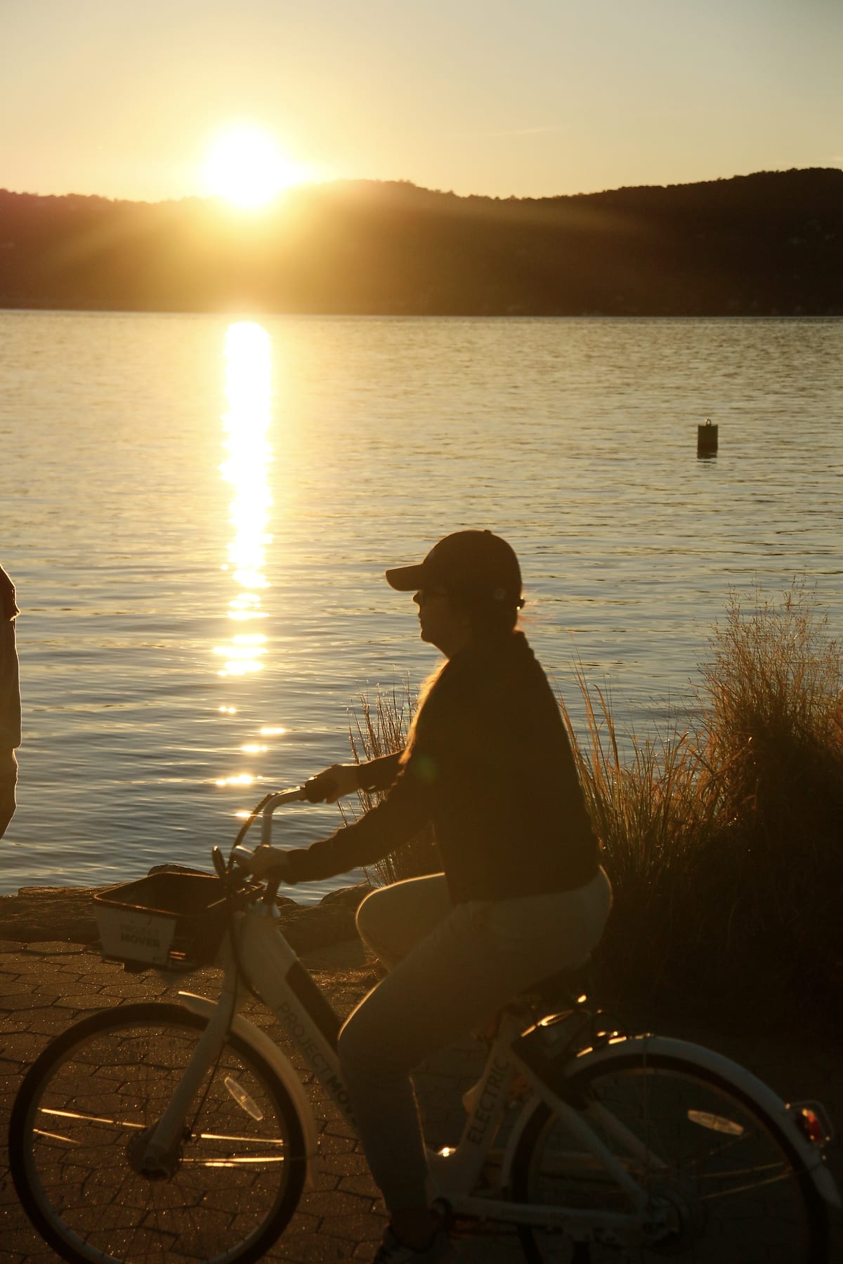 A cyclist in silhouette rides along the river at sunset.