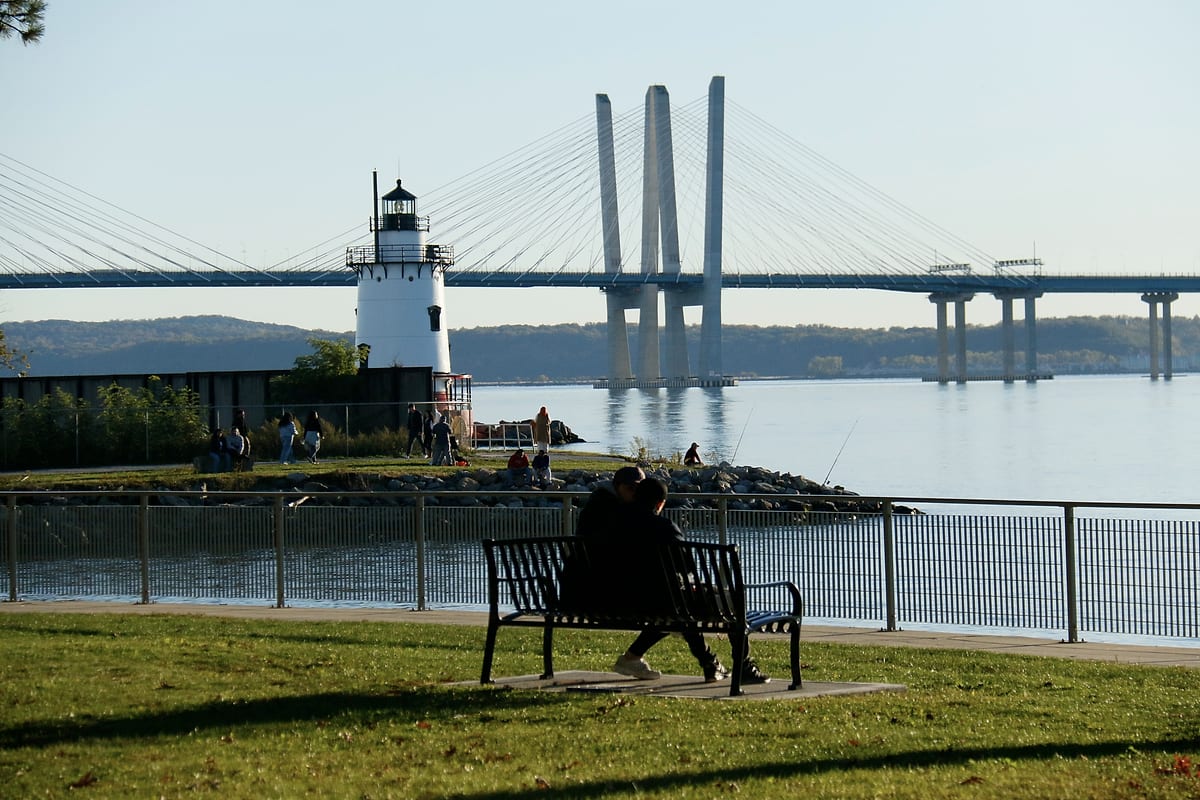 Two people on a bench by the river, the lighthouse and the bridge beyond.
