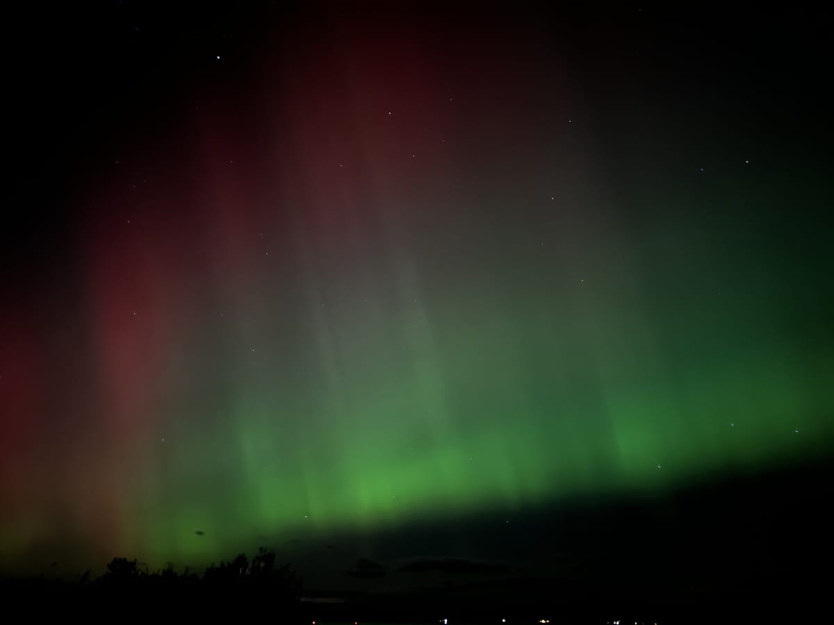 Curtains of green and red aurora above a dark horizon with a few distant lights.