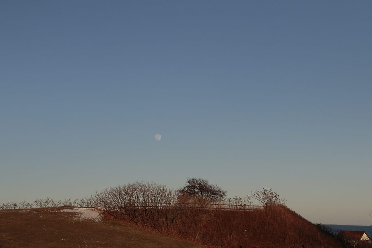 A low hill against the evening sky, the moon small above it.