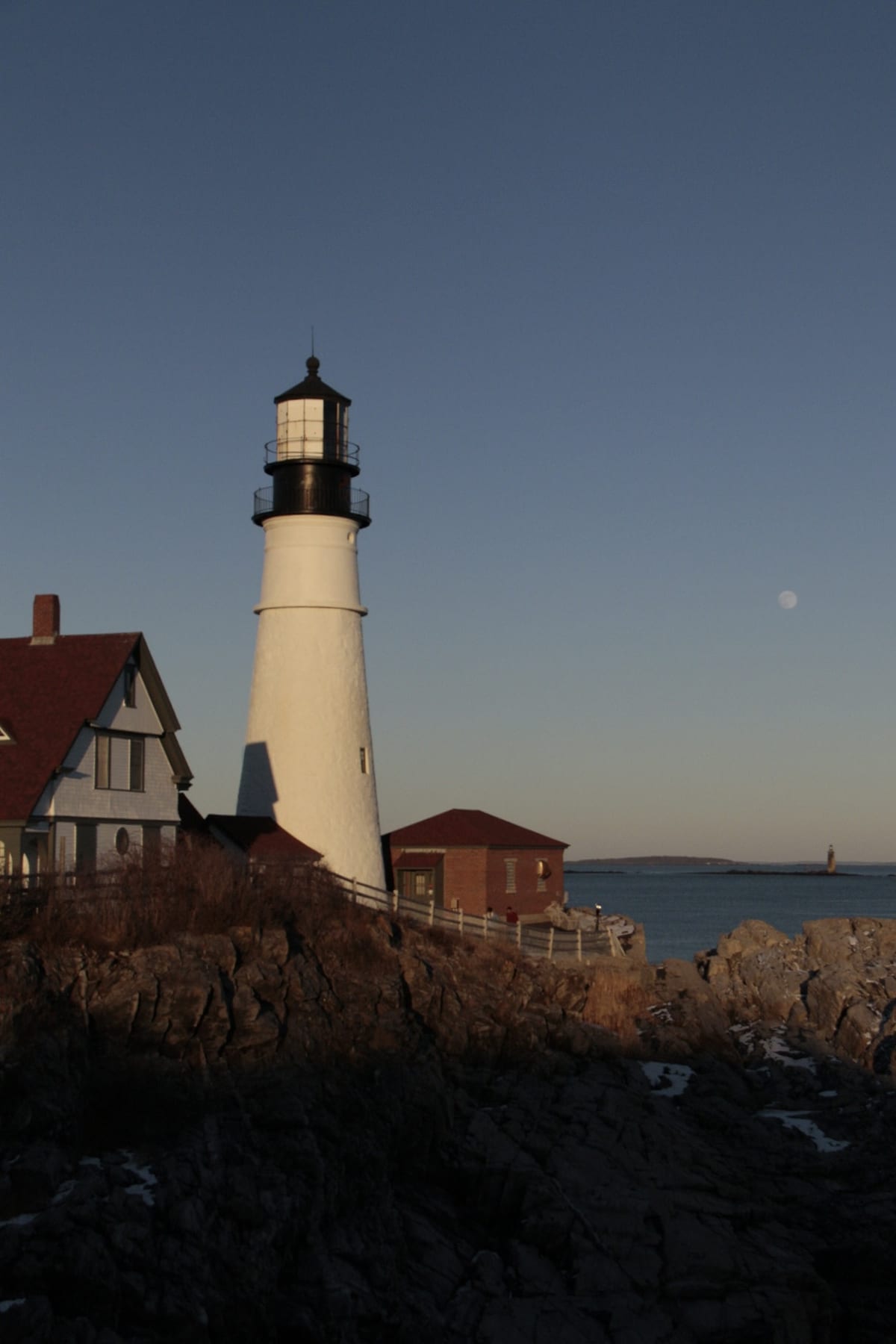 A white lighthouse and its keeper's house on the rocks in late light.